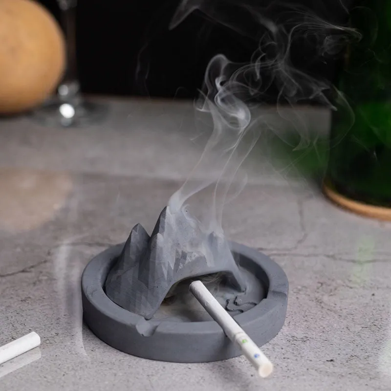 Grey mountain-shaped concrete ash tray holding a cigarette with smoke