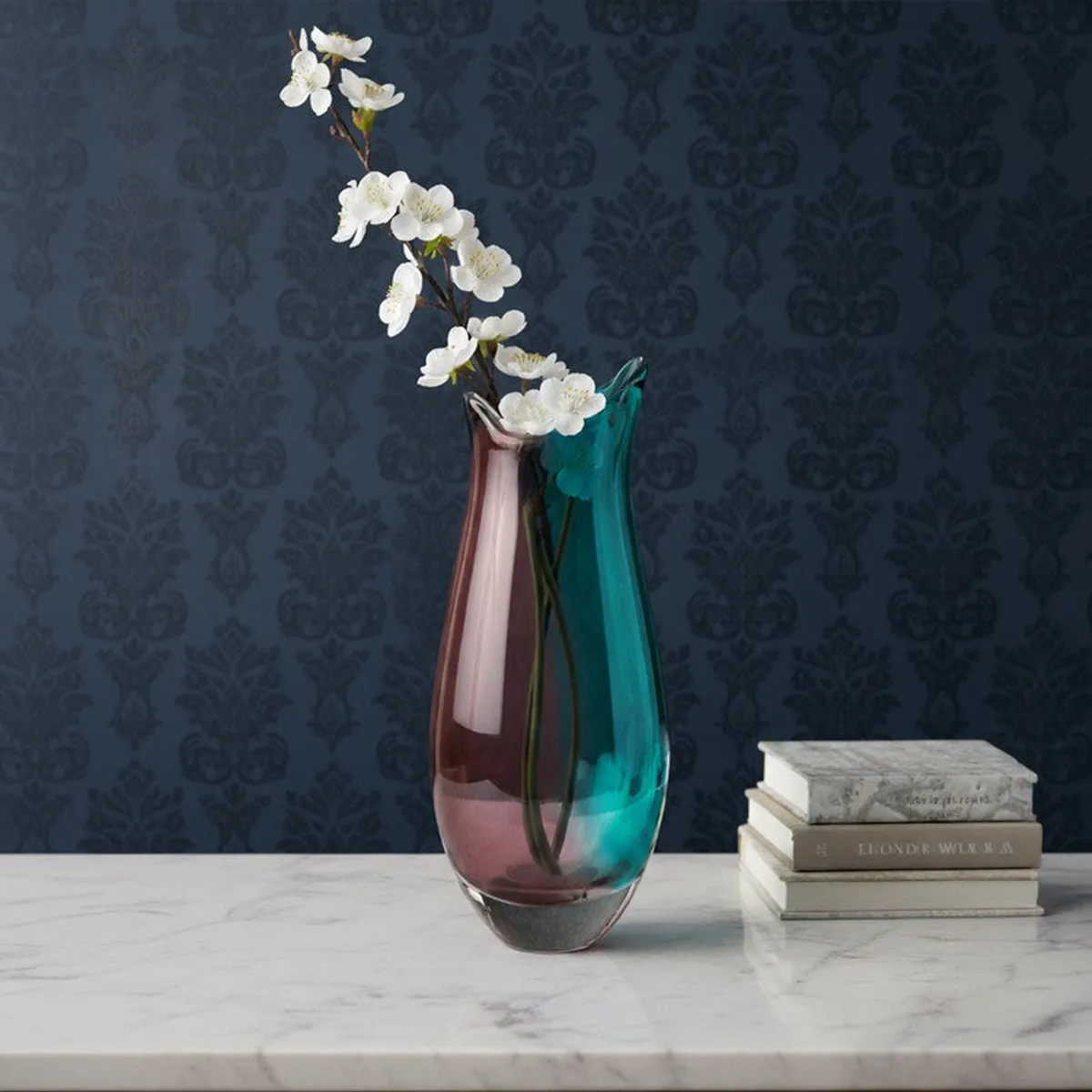 Purple and blue glass vase with white flowers on a marble surface against patterned wallpaper