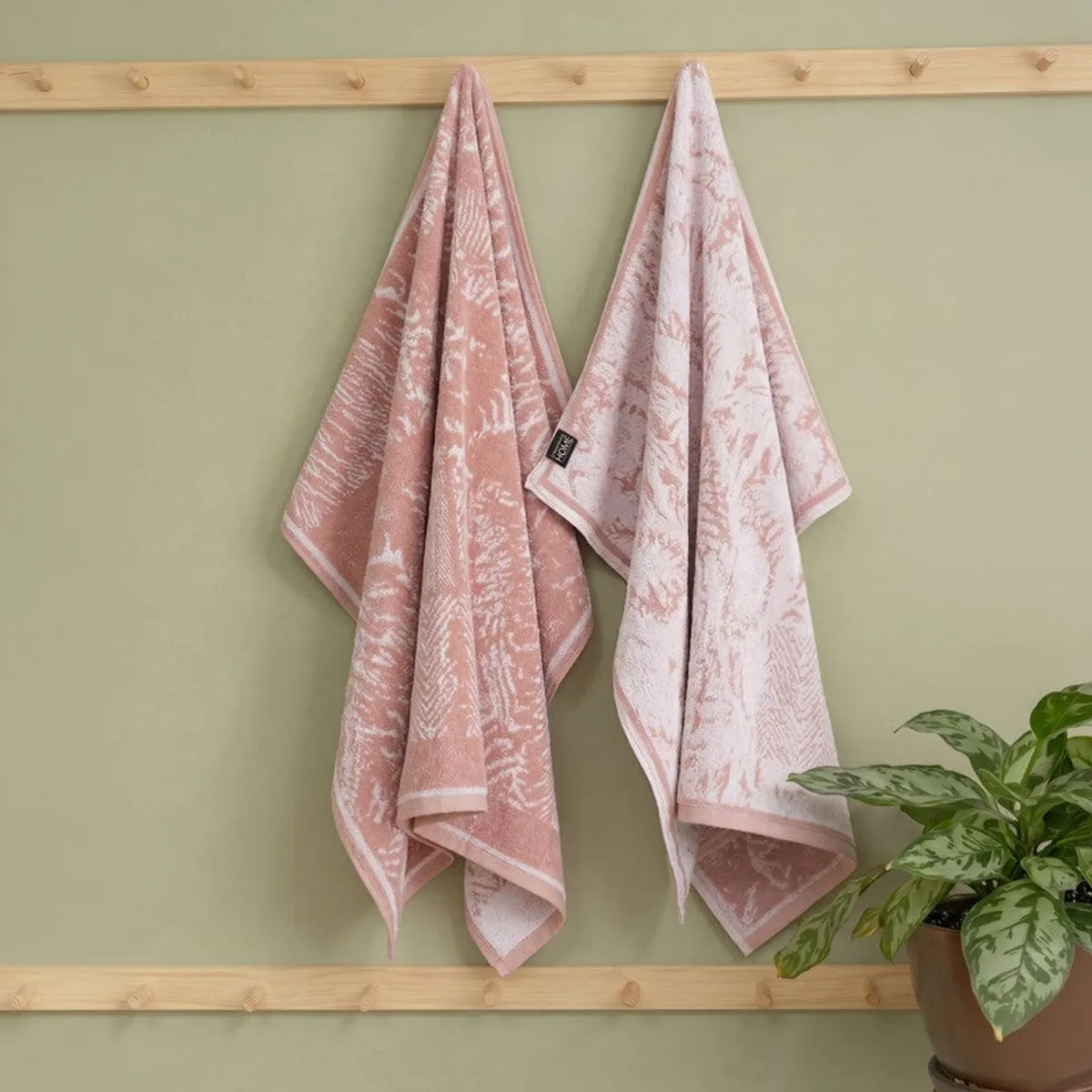 Peach floral bath towel hanging on a wooden peg rail beside a lighter towel and houseplant