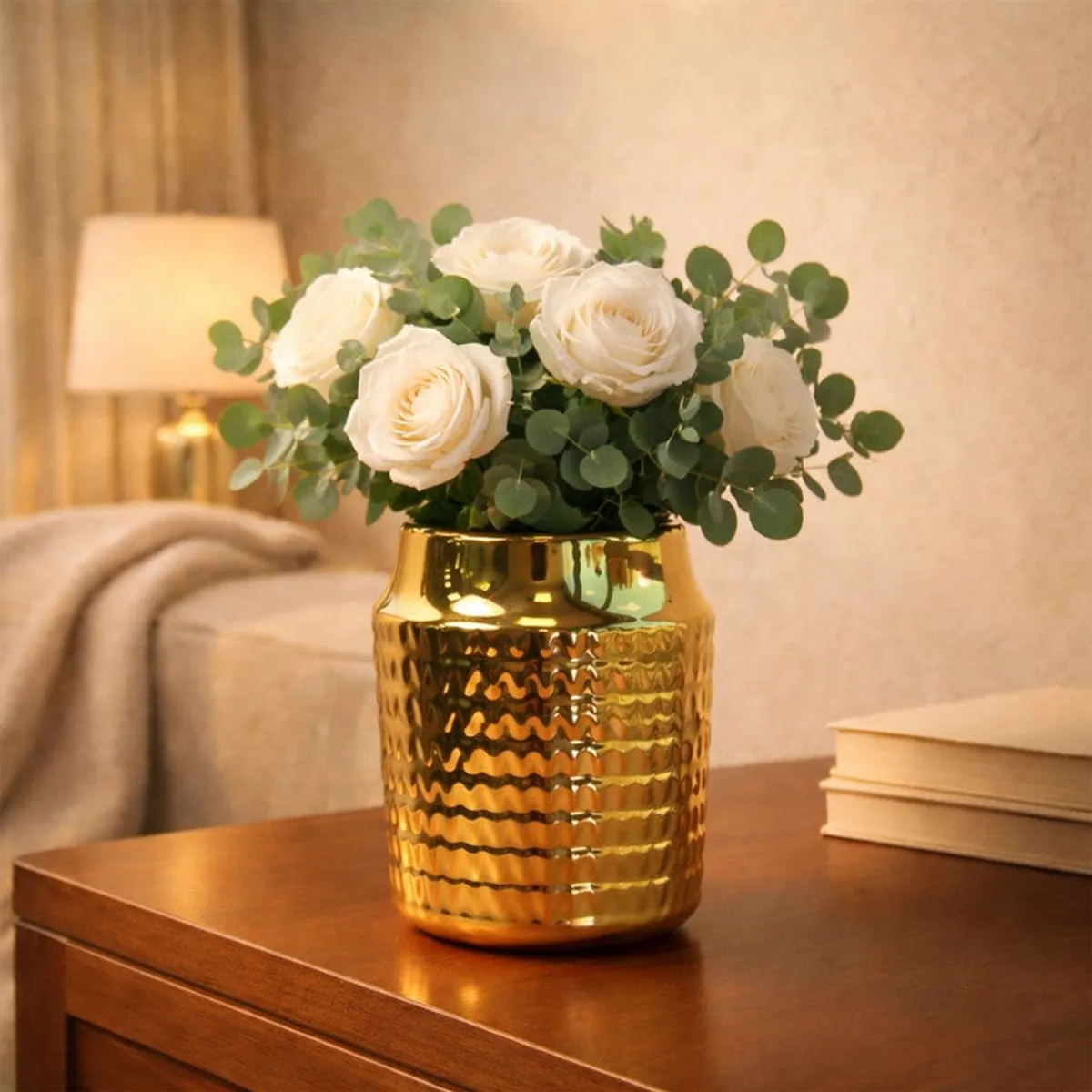 Gold ribbed ceramic vase with white roses and eucalyptus on a wooden side table.