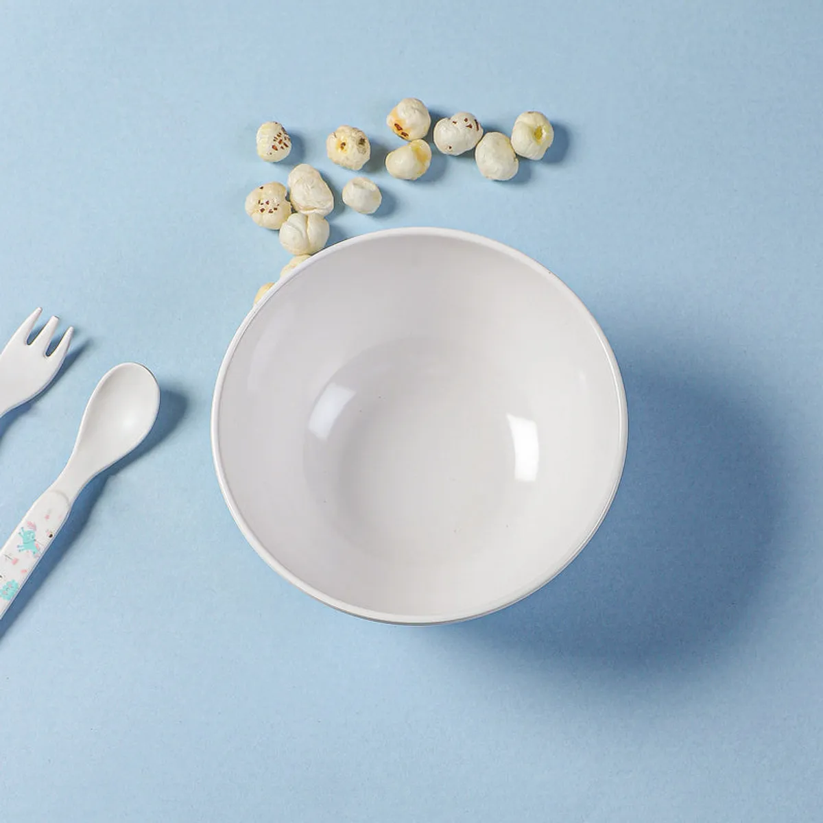 Top view of a white round snack bowl with pastel artwork and snacks nearby