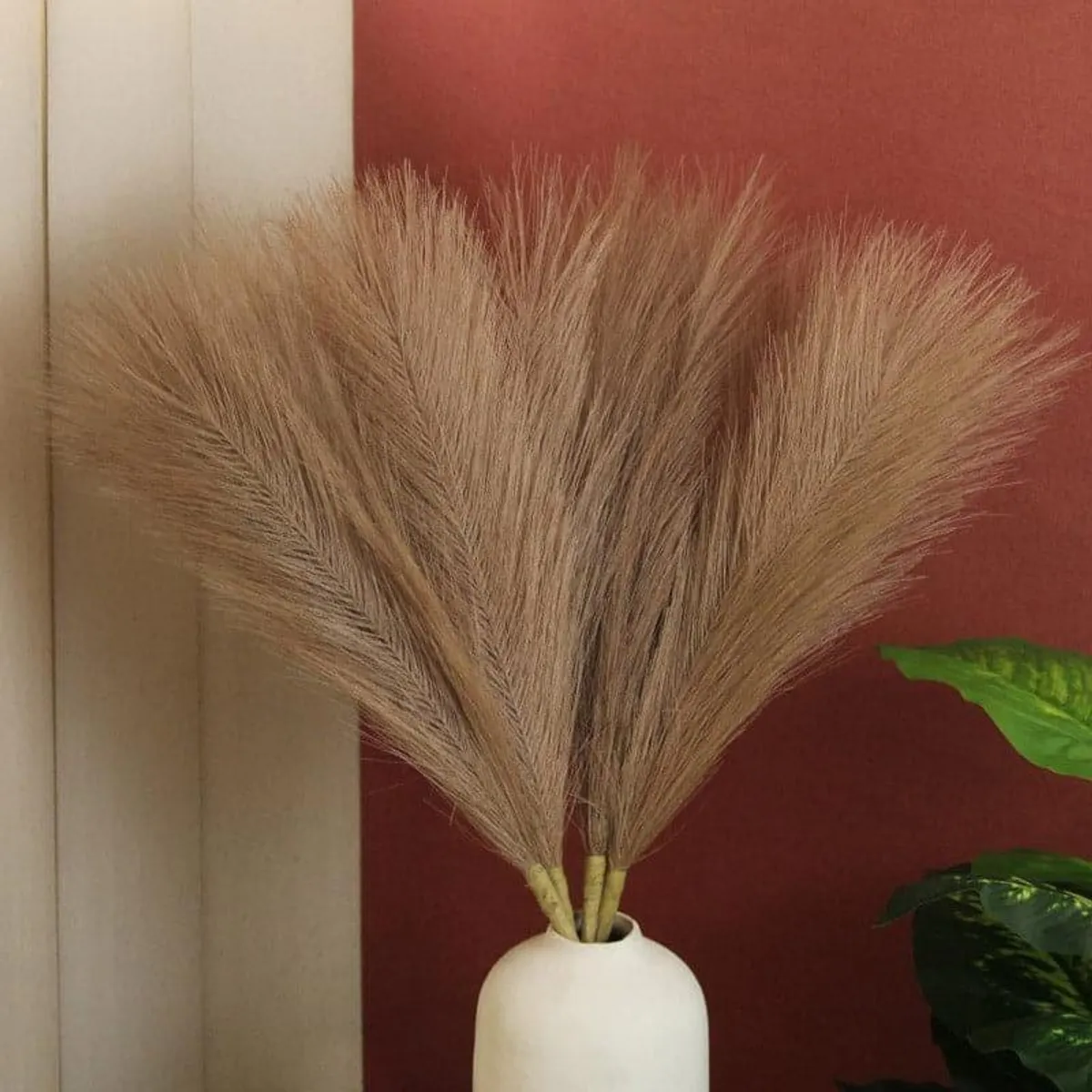Brown feathery faux pampas grass arranged in a white vase against a warm wall