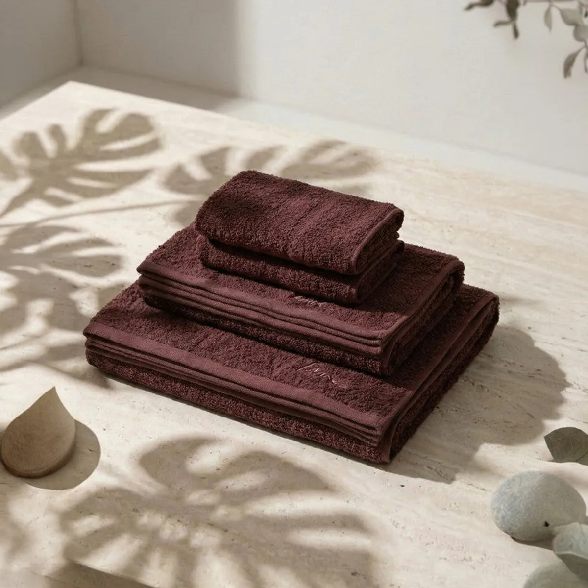 Stack of brown towels arranged on a light stone surface with soft leaf shadows.