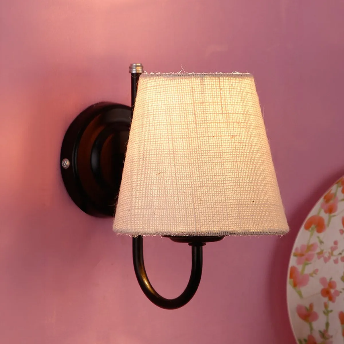 Lit wall lamp with off-white jute shade and black iron base on a pink wall