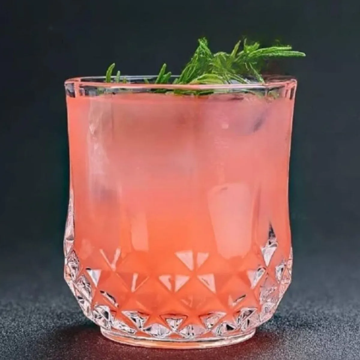 Clear textured whisky glass filled with a pink drink and garnished with herbs