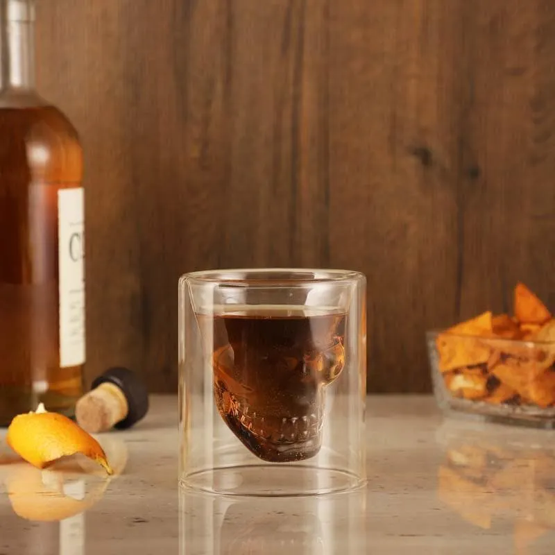 Transparent skull shot glass filled with a dark drink on a bar counter.