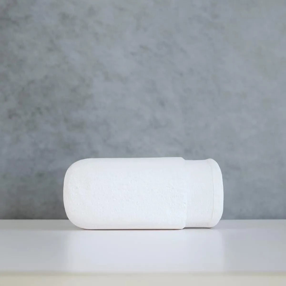Rolled white bath towel displayed on a light surface against a grey backdrop