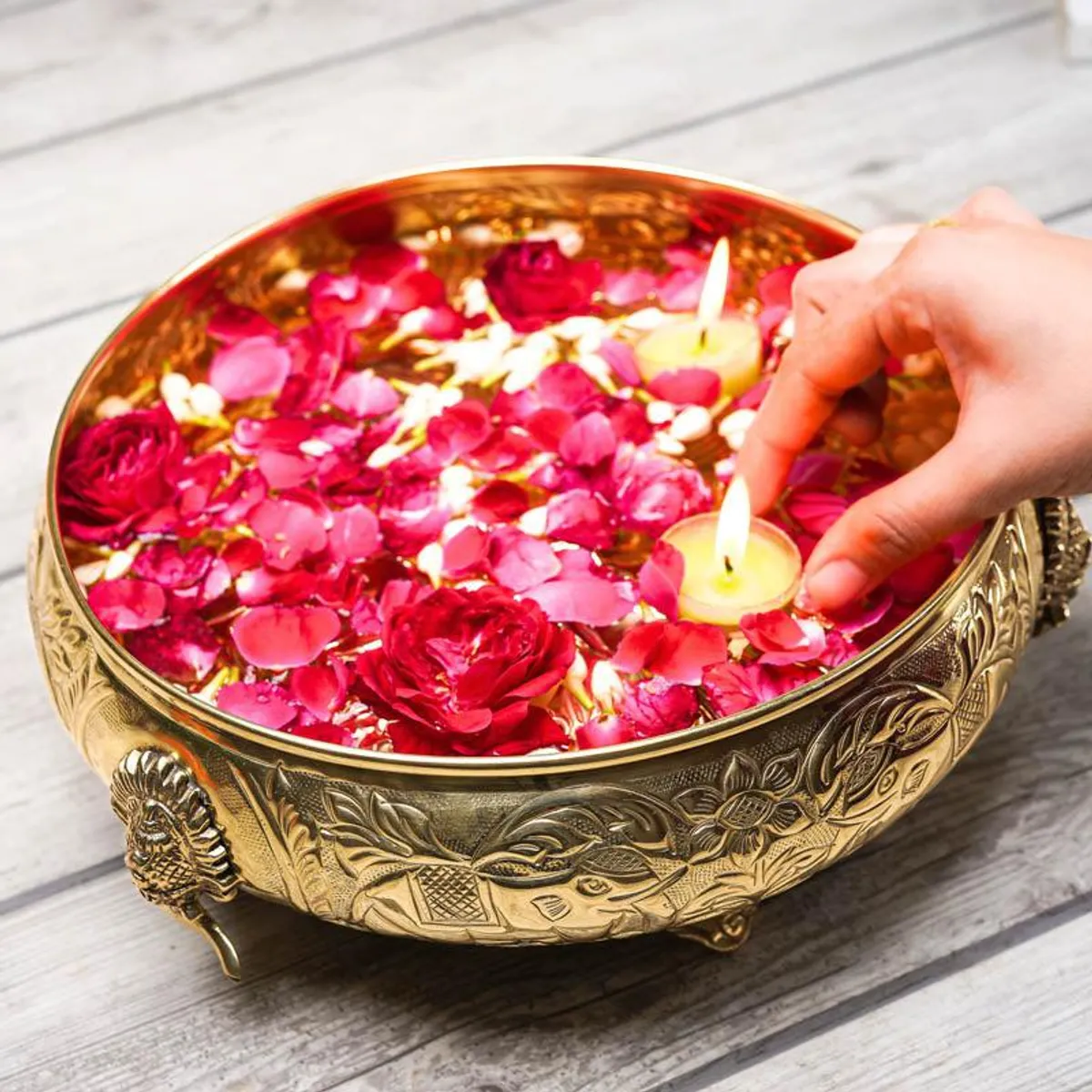 A hand places a floating candle among rose petals in the gold urli.