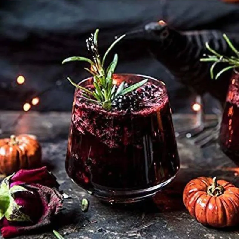 Clear tumbler holding a dark cocktail with rosemary garnish in a moody setting.