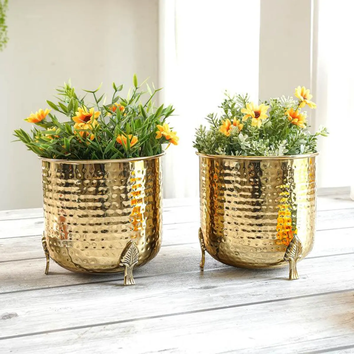 Two gold hammered metal planters with leaf-shaped legs holding faux flowers
