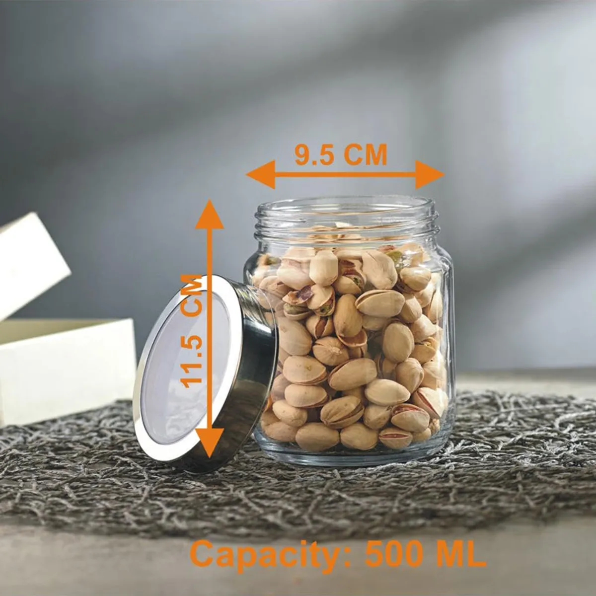 Transparent glass storage jar filled with pistachios and its metallic lid beside it.