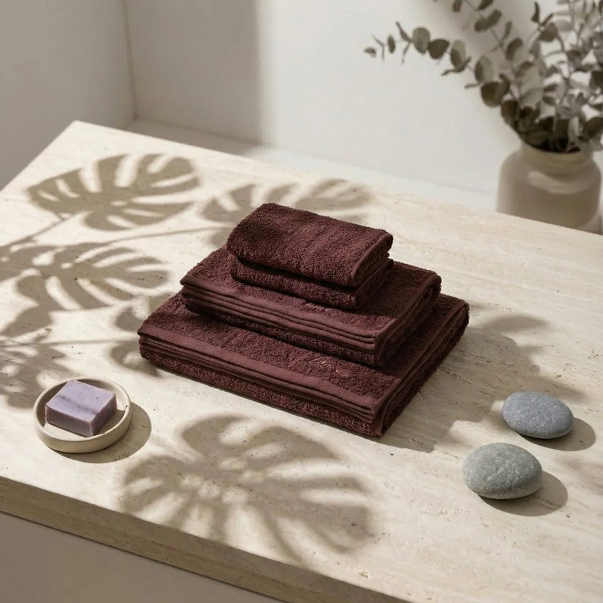 Folded brown towel set on a light surface with soap, stones and vase in view.