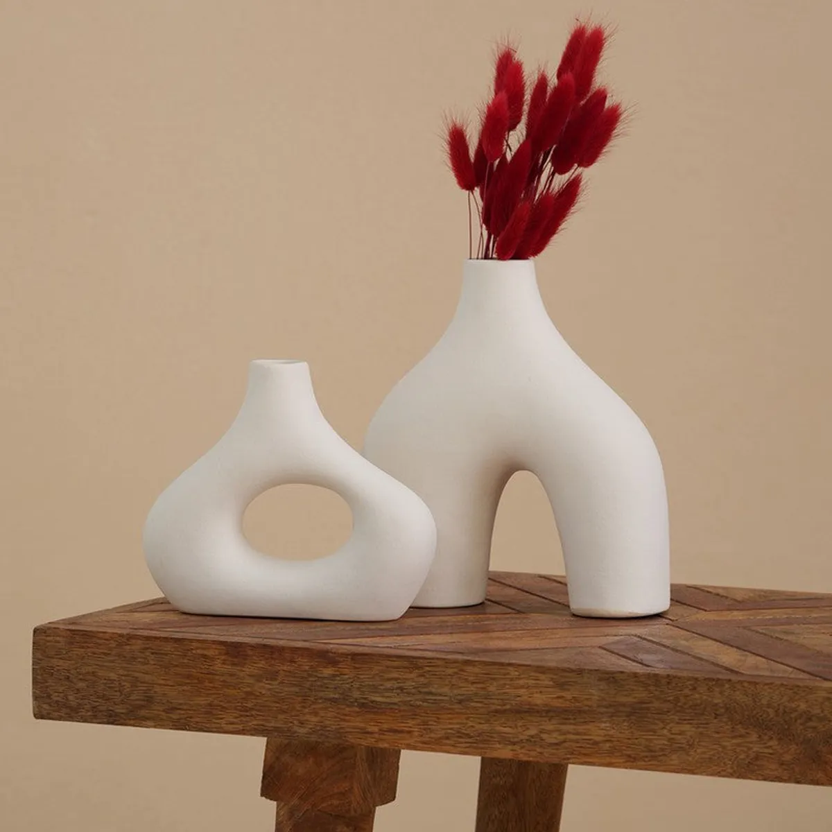 Two white hollow-out ceramic vases styled on a wooden table with red dried stems.