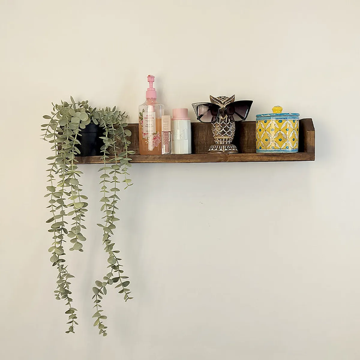 Brown wooden wall shelf displaying a trailing plant, toiletries, an owl figurine and a decorative box.