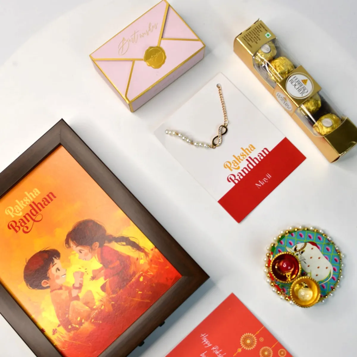 Rakhi, chocolate, greeting card, framed artwork and decorated roli thali arranged on a white surface.