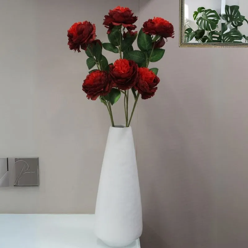 Red artificial rose flower bunches displayed in a white vase against a grey wall.