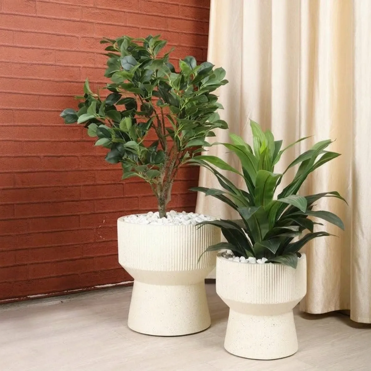 Two off-white hourglass planters displayed with indoor plants near curtains and a brick wall.