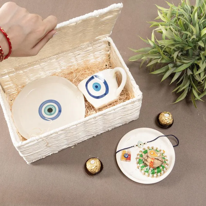 Open white hamper box with evil eye ceramic cup and saucer beside a rakhi platter