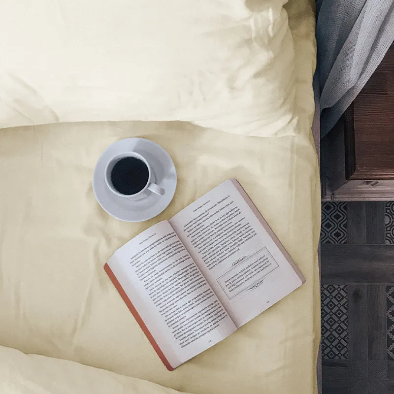 Close-up top view of an ivory bedsheet with a pillow, cup and open book