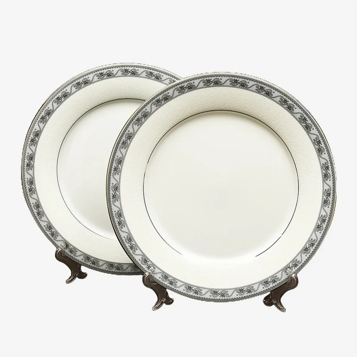Two white porcelain dinner plates with ornate borders on display stands.