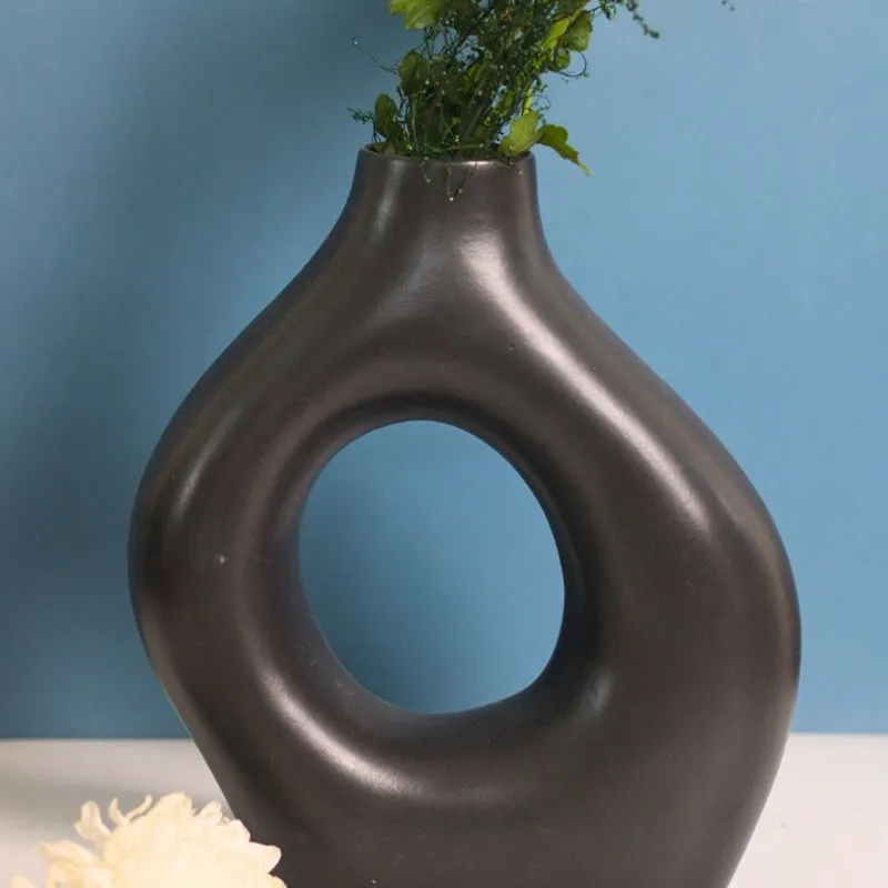 Black donut-shaped ceramic vase with foliage against a blue background