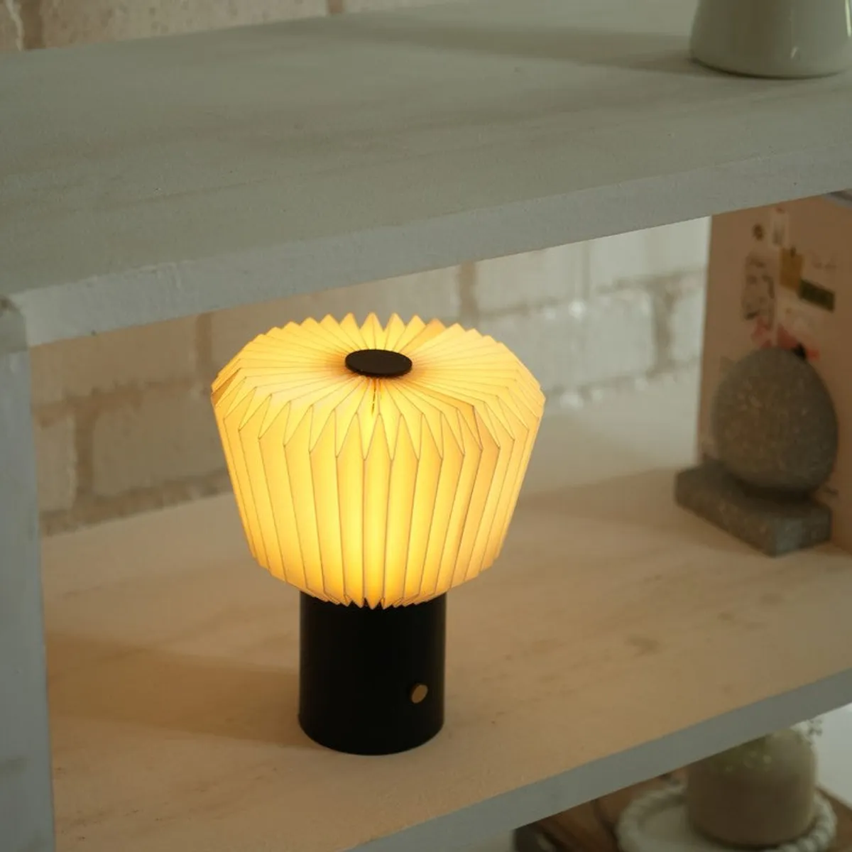 Glowing pleated table lamp displayed on a shelf with a black cylindrical base