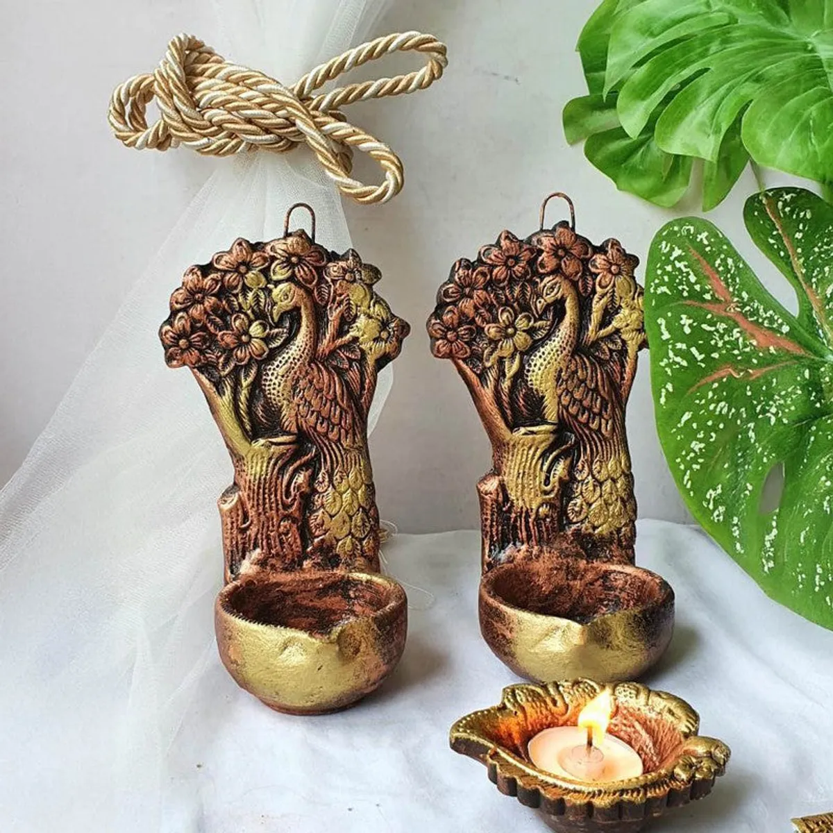 Two peacock-shaped diyas displayed against white fabric and green leaves