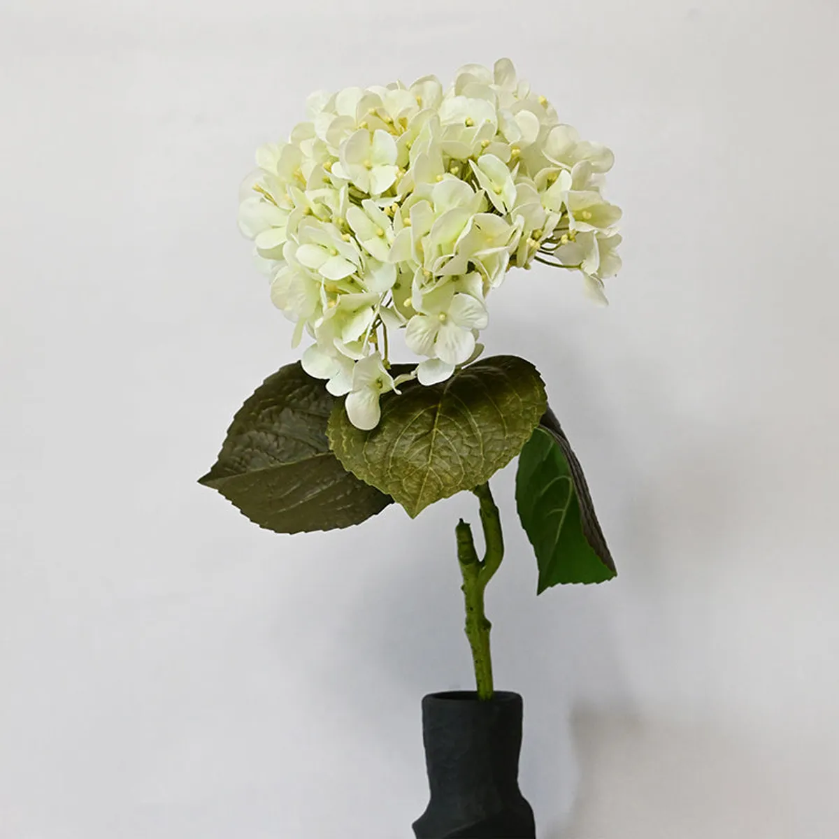 White artificial hydrangea flower and leaves shown with its stem in a vase.