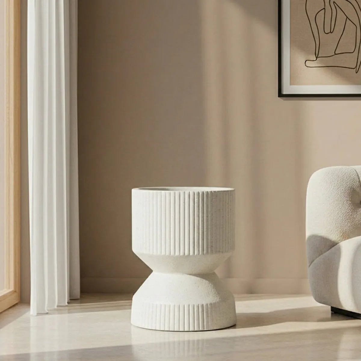 Off-white hourglass planter shown empty beside a chair in a modern room.