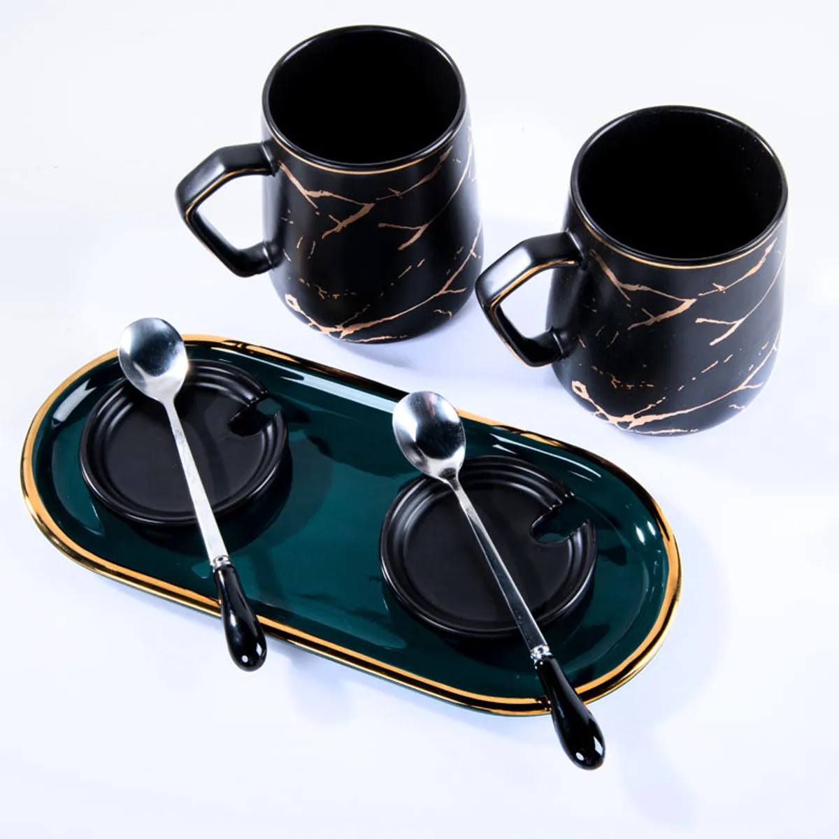 Two black mugs, spoons and a deep green tray arranged on a white background