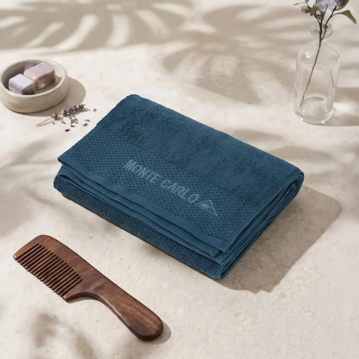 Folded blue bath towel with Monte Carlo branding beside soap and a wooden comb