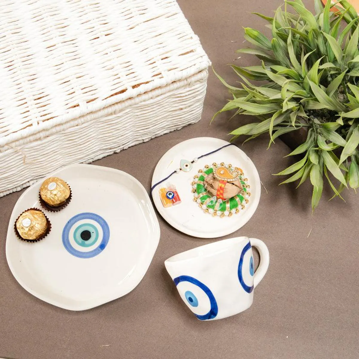 Ceramic evil eye saucer, cup, rakhi platter and chocolates arranged beside a white hamper box