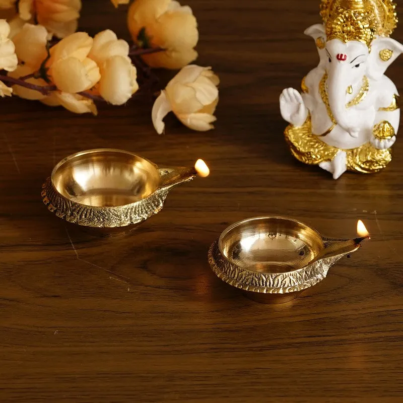Two lit gold brass diyas styled on a wooden table beside a Ganesha idol