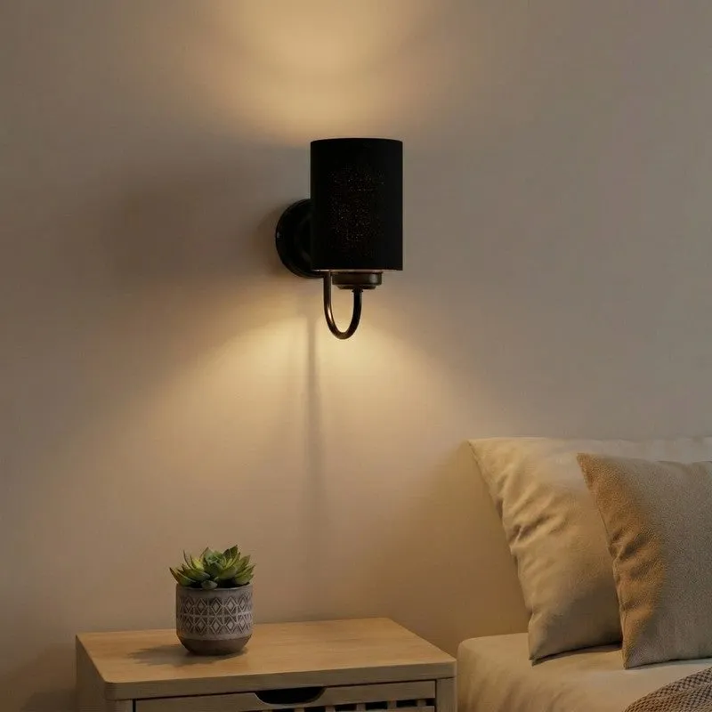Matte black cylindrical wall lamp mounted beside a bed, casting warm upward light