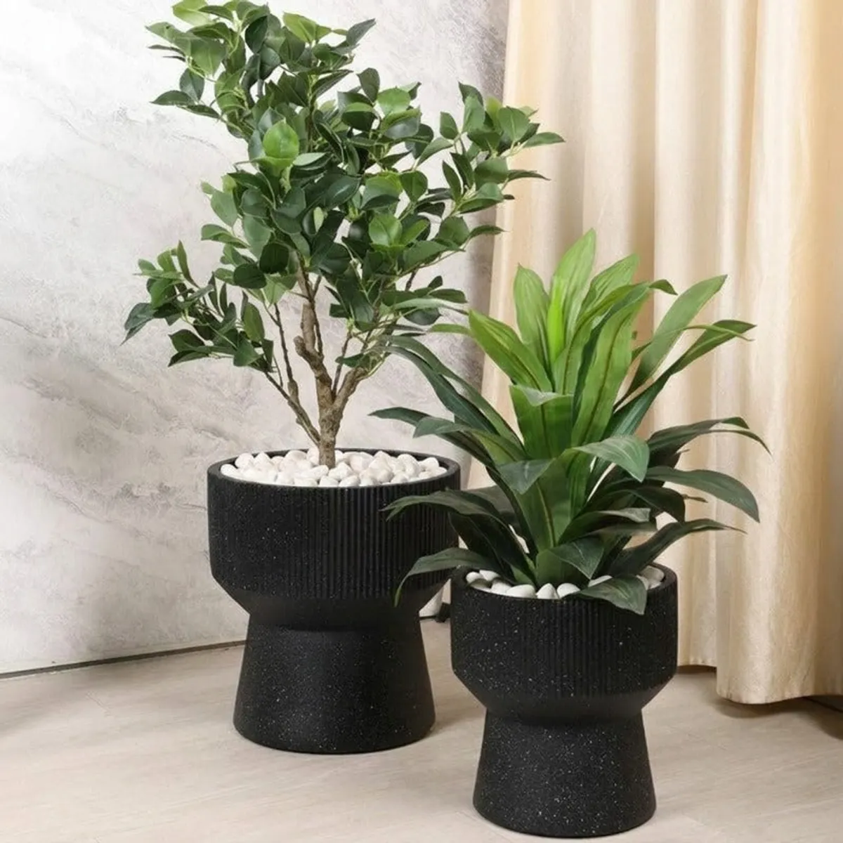 Two black ribbed pedestal planters displayed with green plants indoors.
