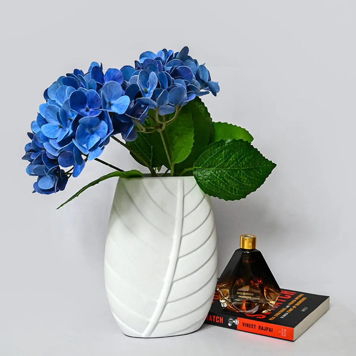 White leaf-pattern vase styled with blue hydrangeas beside a perfume bottle and book