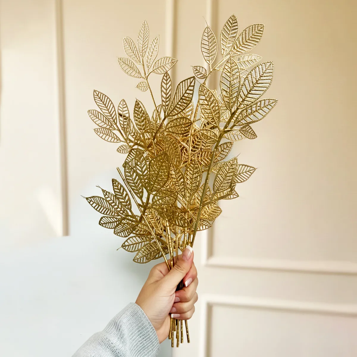 Hand holding two golden artificial leaf stems against a light background