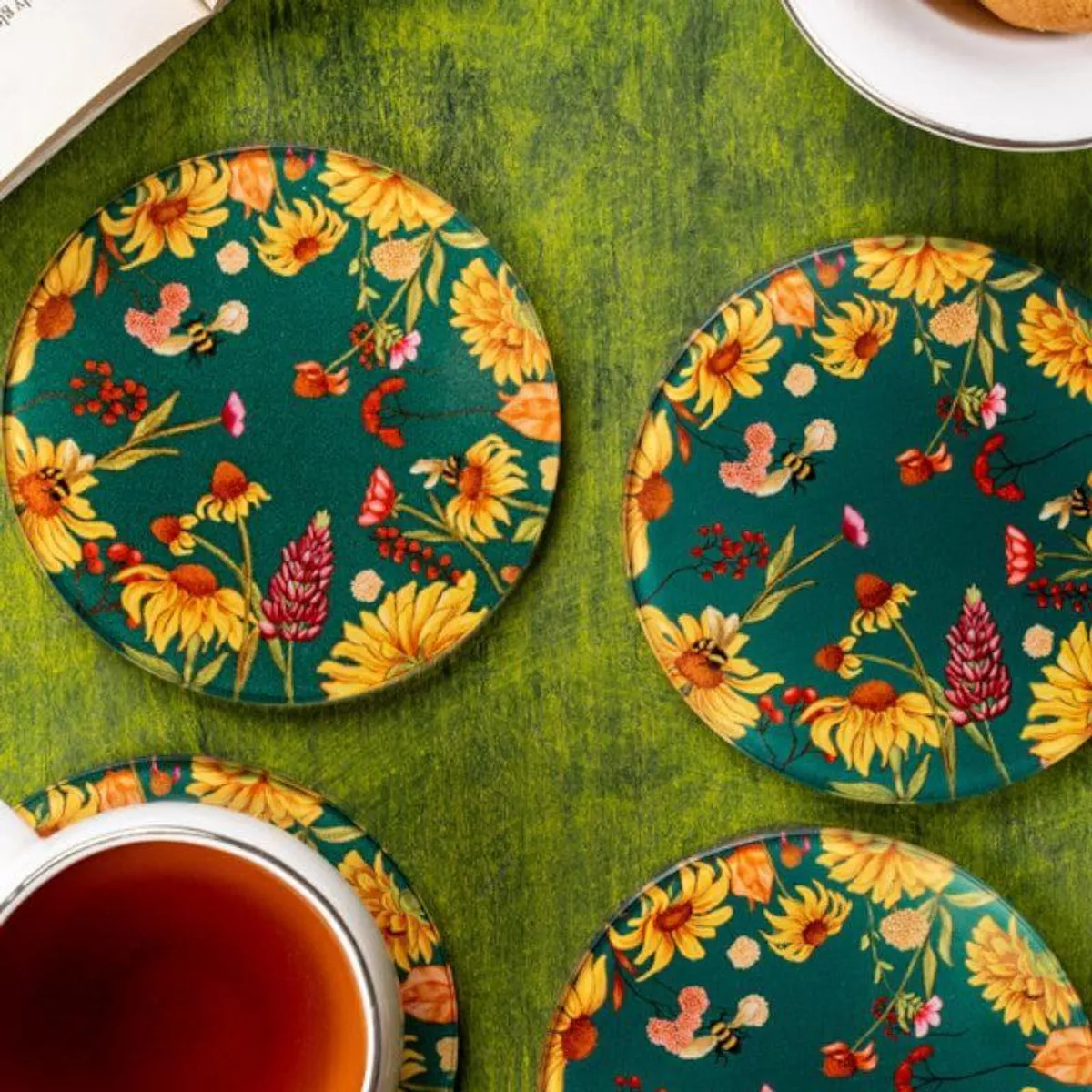 Three floral round coasters on a green tabletop with a teacup, cookies and an open book.