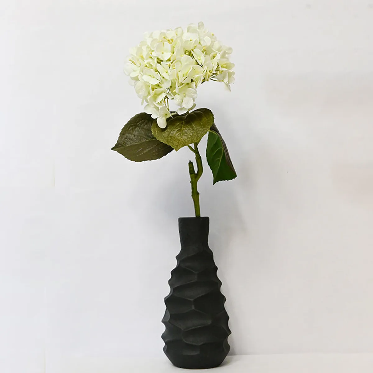 White artificial hydrangea stick displayed in a tall black textured vase.
