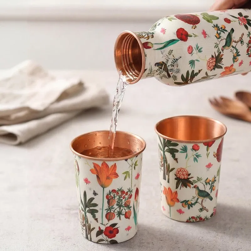 Floral copper bottle pouring water into one of two matching copper tumblers