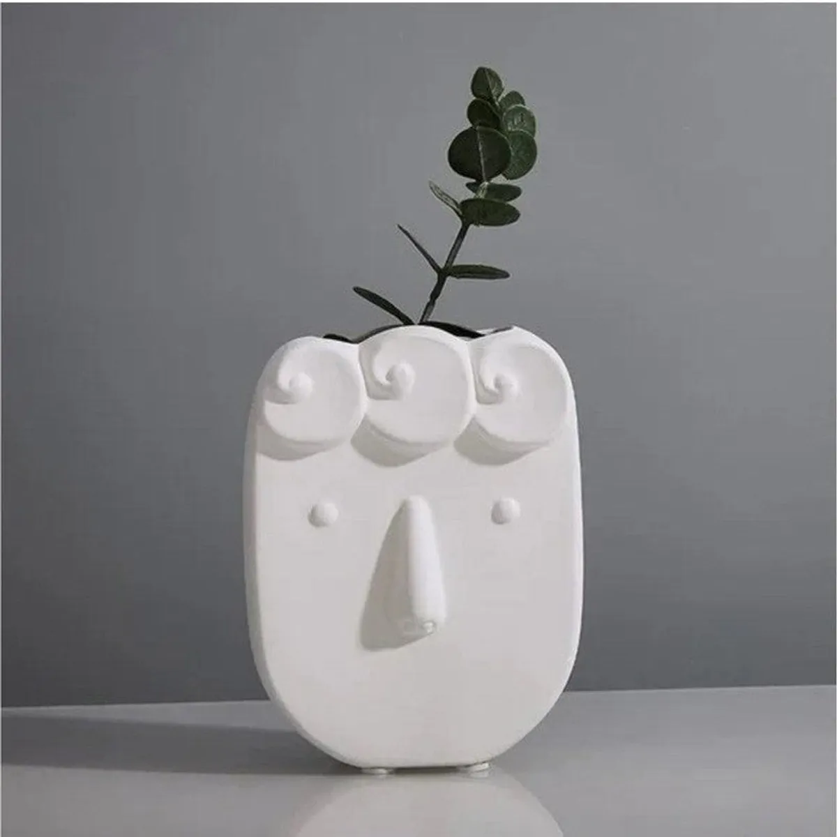 White face-shaped vase with curled hair detail holding a green stem against a grey background