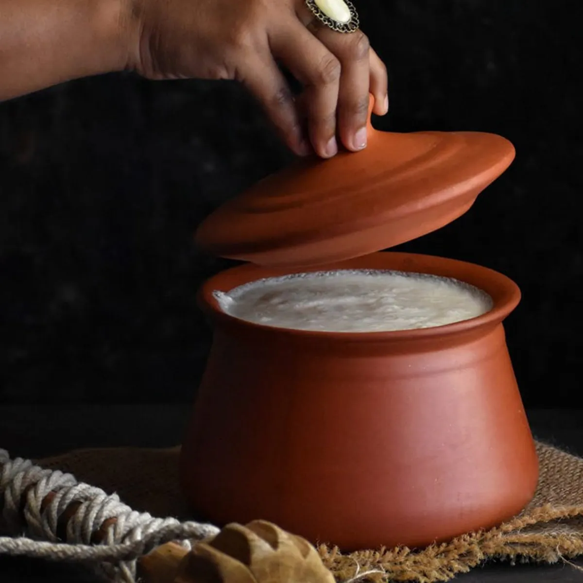 Hand lifting the lid of a brown terracotta handi filled with food