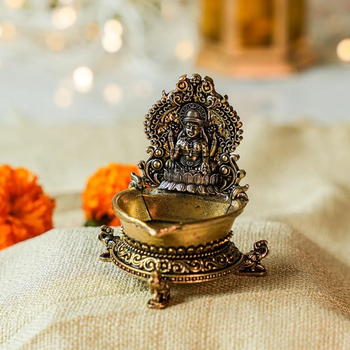 Gold-toned Lakshmi diya displayed on a textured fabric surface.