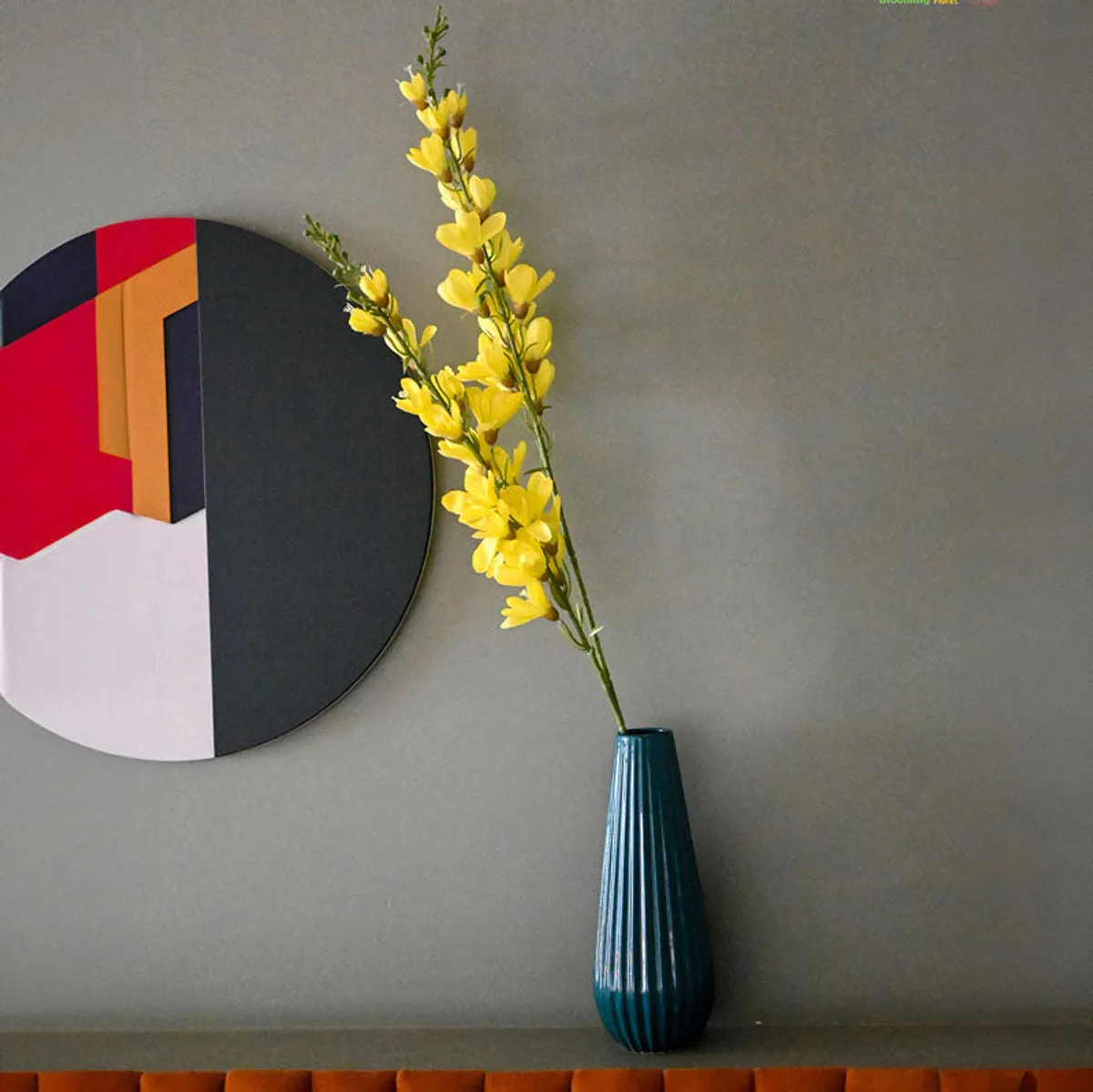 Yellow artificial flower stick displayed in a tall teal vase beside geometric wall art.