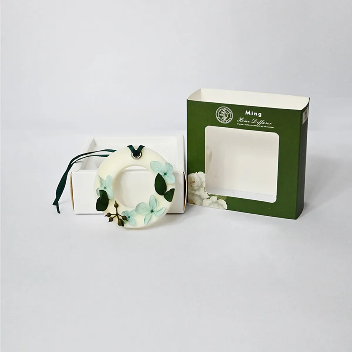 Floral round wax melt with ribbon placed in front of open green packaging box