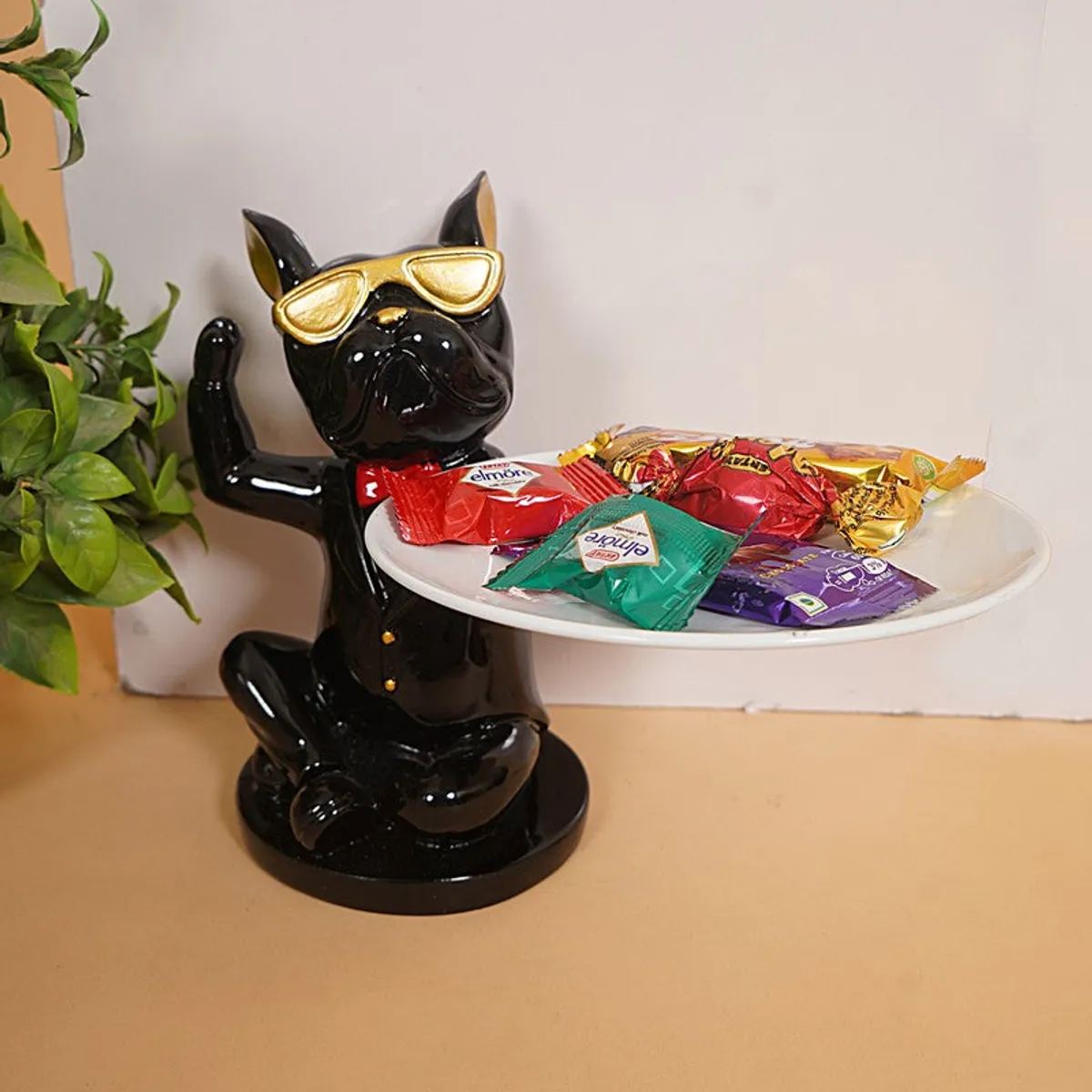 Black tray-holding figurine styled beside a potted plant with wrapped sweets on the tray