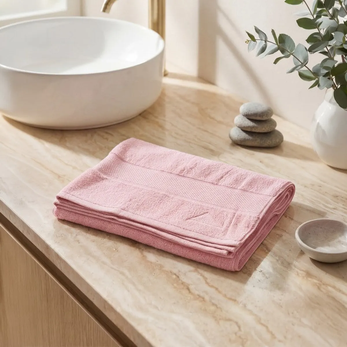Folded light pink bath towel styled beside a basin and decorative stones