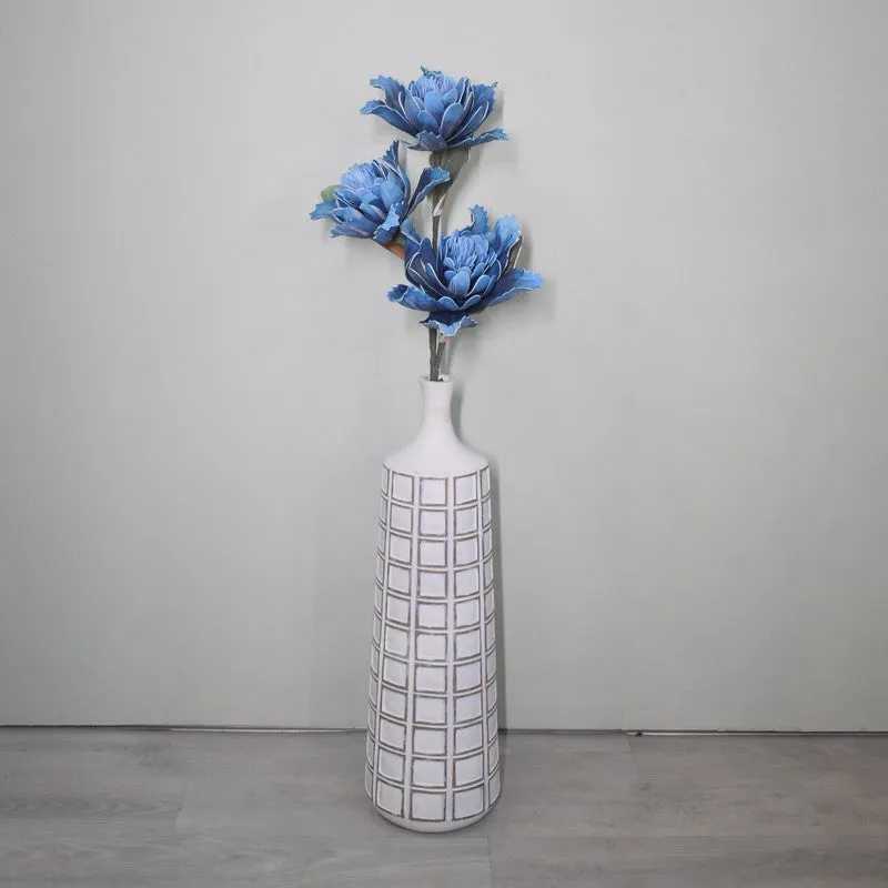 Blue artificial dahlia stem standing in a tall white vase against a plain wall.