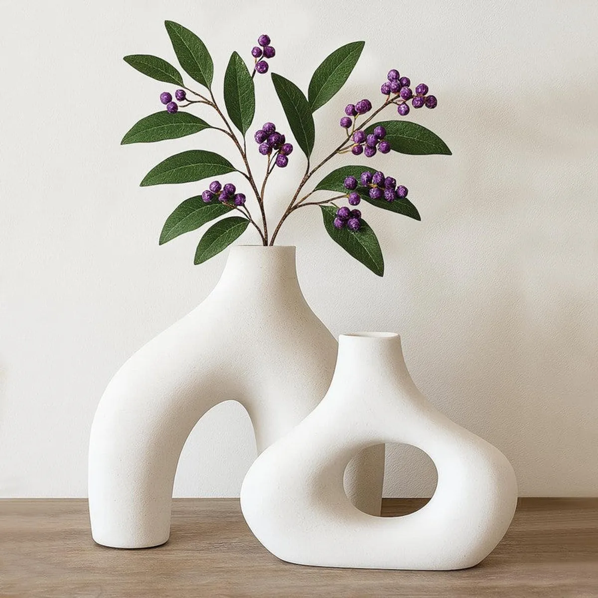 Two matte white vases on a wooden surface, with leafy purple berry stems in the taller vase.