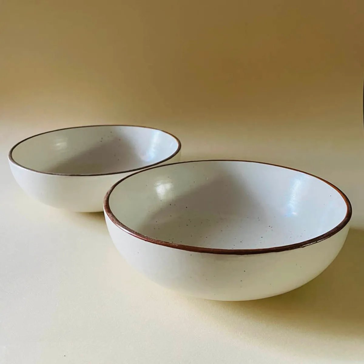 Two beige bowls with brown rims shown separately on a warm neutral surface.